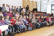 Große Gruppe von Grundschulkindern, die in einem Chor singen und dazu Gesten mit den Händen machen.