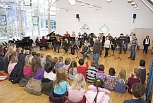 Ein Orchester aus etwa 10 Erwachsenen spielt vor eine größeren Gruppe von Kindern.