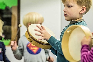 Zwei Vorschulkinder spielen auf Rahmentrommeln, ein weiteres schwingt im Hintergrund Schlegel für ein Xylophon