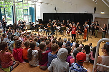 Ein Orchester aus etwa 10 Erwachsenen spielt vor eine größeren Gruppe von Kindern.