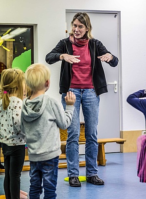 Musiklehrerin zeigt Tanzgesten mit den Armen, vor ihr stehen Vorschulkinder, die die Bewegungen nachahmen