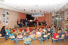 5 Erwachsene spielen von einer kleinen Bühne mit Akkordeon, Flügel und Keyboard vor einer größeren Gruppe Grundschulkindern.